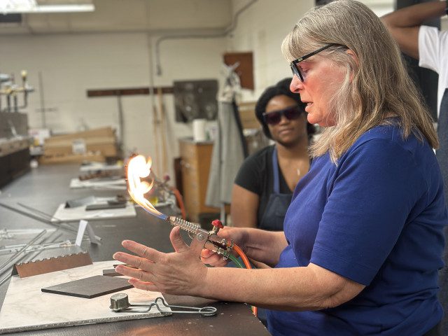 Woman wearing blue shirt and glasses blows glass.